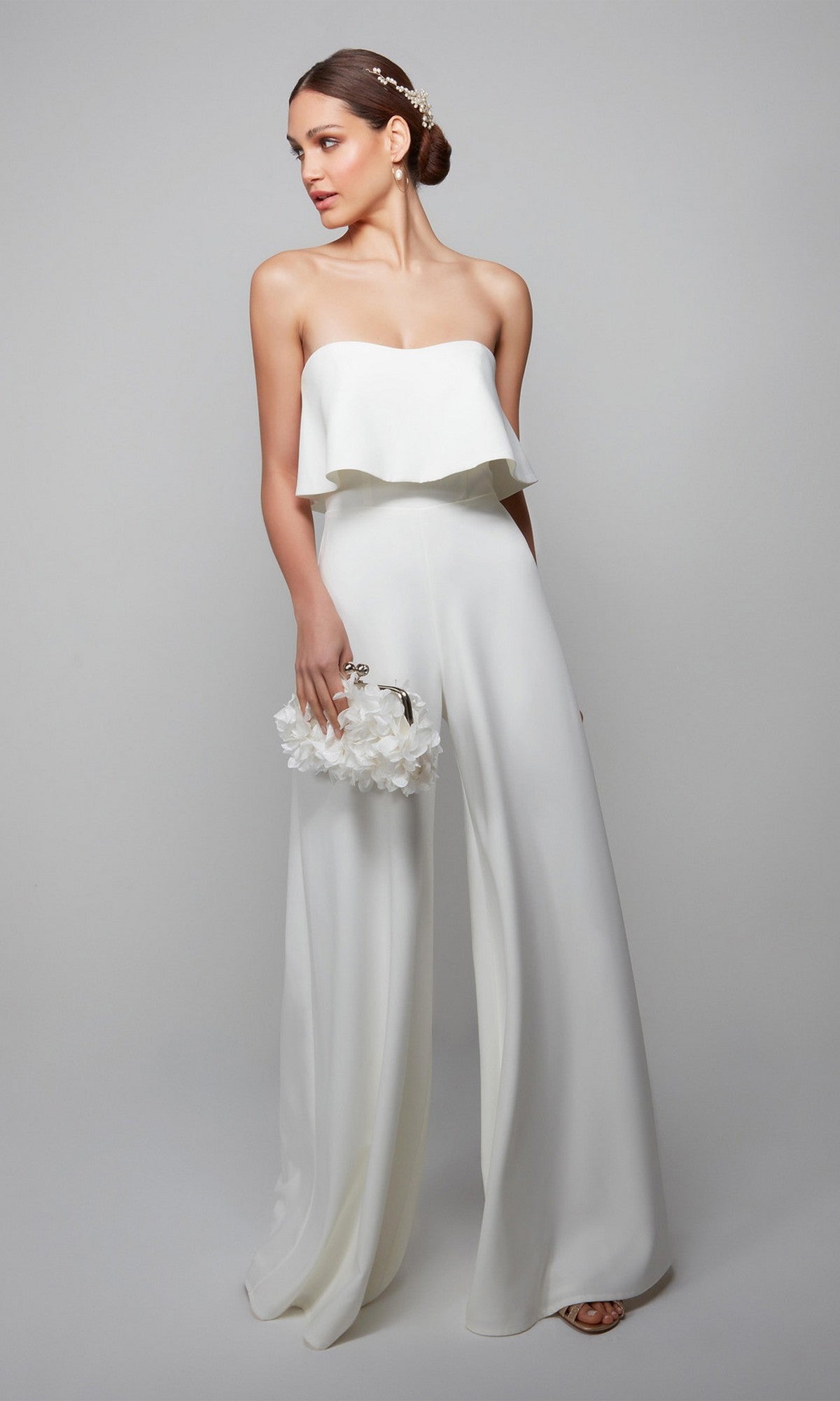 A woman in the Long Formal Jumpsuit: Alyce 70015, featuring wide-leg pants, holds a white floral clutch and has her hair styled in an updo adorned with a hair accessory.
